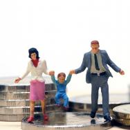 Two miniature figures lift their child up while standing on a pile of coins