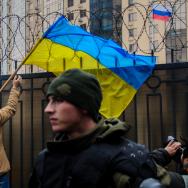 Man holds a Ukraine flag near barbed wire, with a Russian flag visible in the background