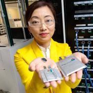 Prof Shirley Meng in Argonne Lab standing in front of a server bank. She's wearing goggles, a yellow lab coat, and holding two lithium batteries in her outstretched hands. 