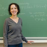 Prof. Barber stands in front of a chalkboard with equations, looking at the camera