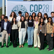 A group of 18 people standing in two rows pose in front of a sign that says COP28
