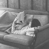 Black and white image of a woman lying down on a couch wrapp while reading a book 
