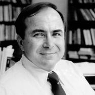 Black and white photo of Mazenko in a white shirt and tie looking at camera in front of bookcases