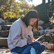 A college student sits on stone steps outside while sketching or writing on a piece of paper.