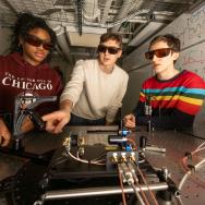 Three students in dark optic glasses at the controls of a large board with scientific equipment.