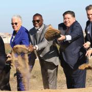 Photo of a row of people in formal dress standing side by side on a sunny day, holding shovels and tossing a shovelful of dirt.