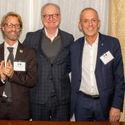 Three smiling men in suits stand above a low table.