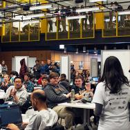 Two people speaking to seated students at a hackathon